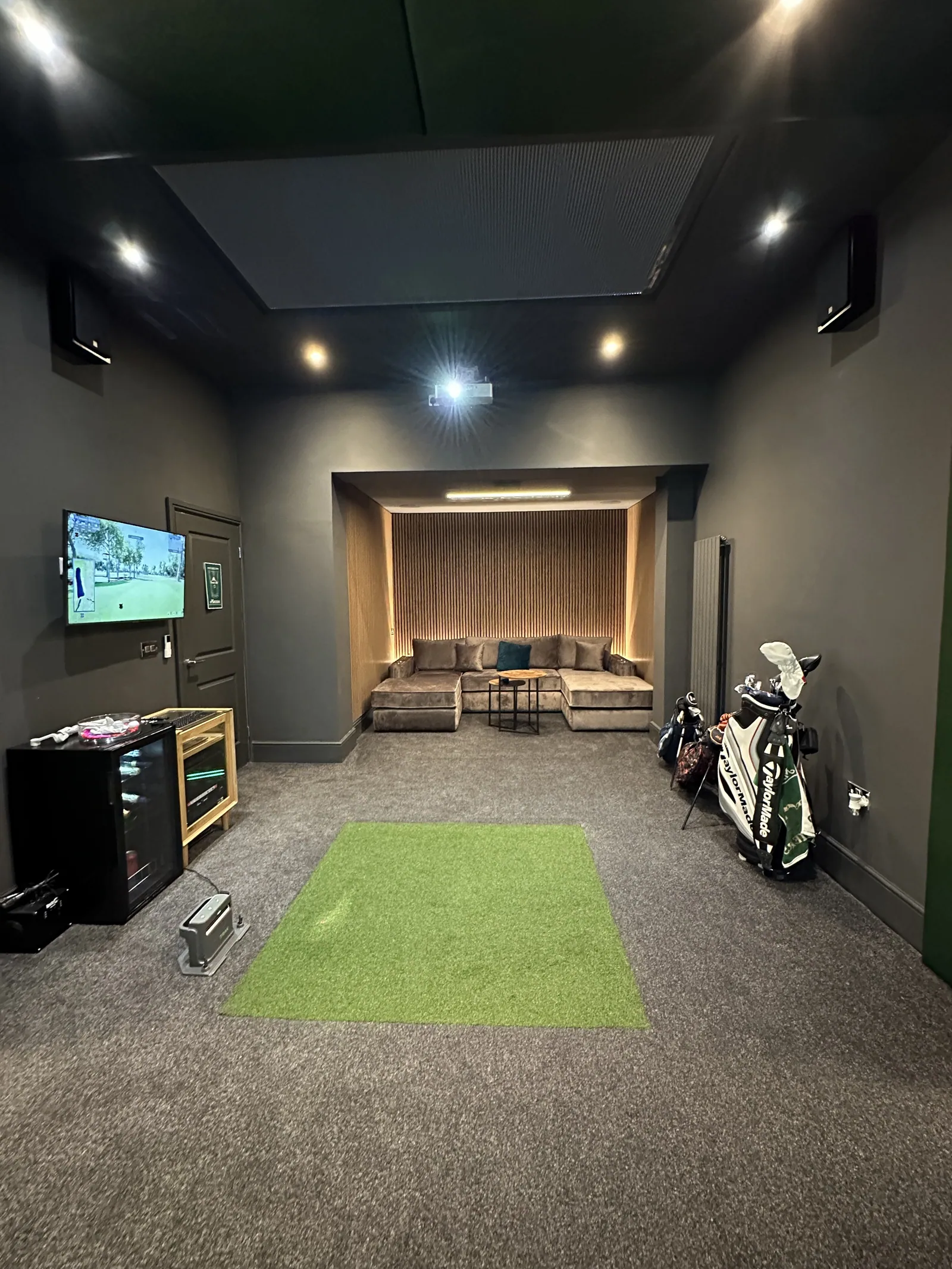 A finished simulator room with a lit screen, turf mat and a lounge area behind