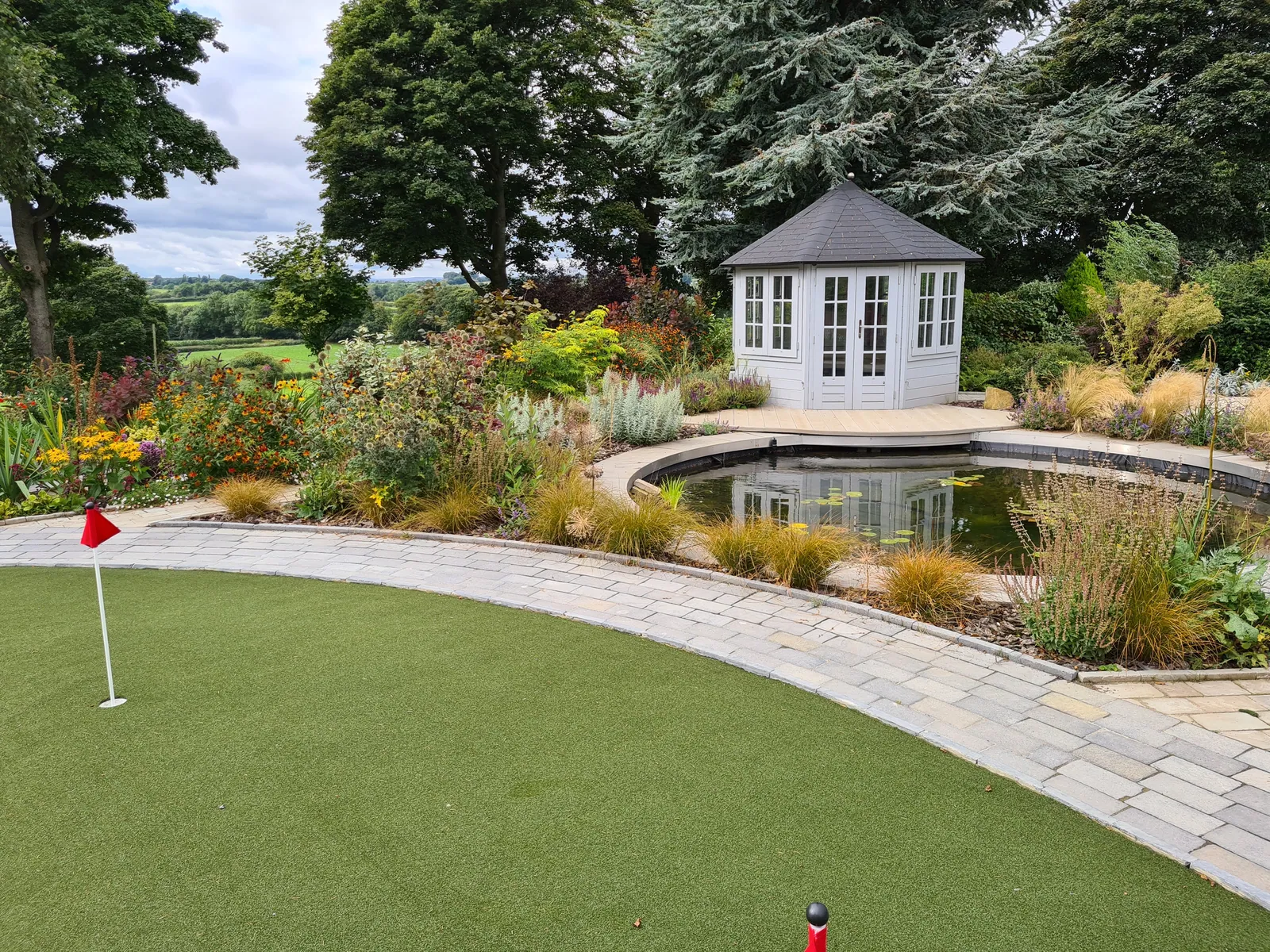A putting green curving around a pond and planting, with a summerhouse behind