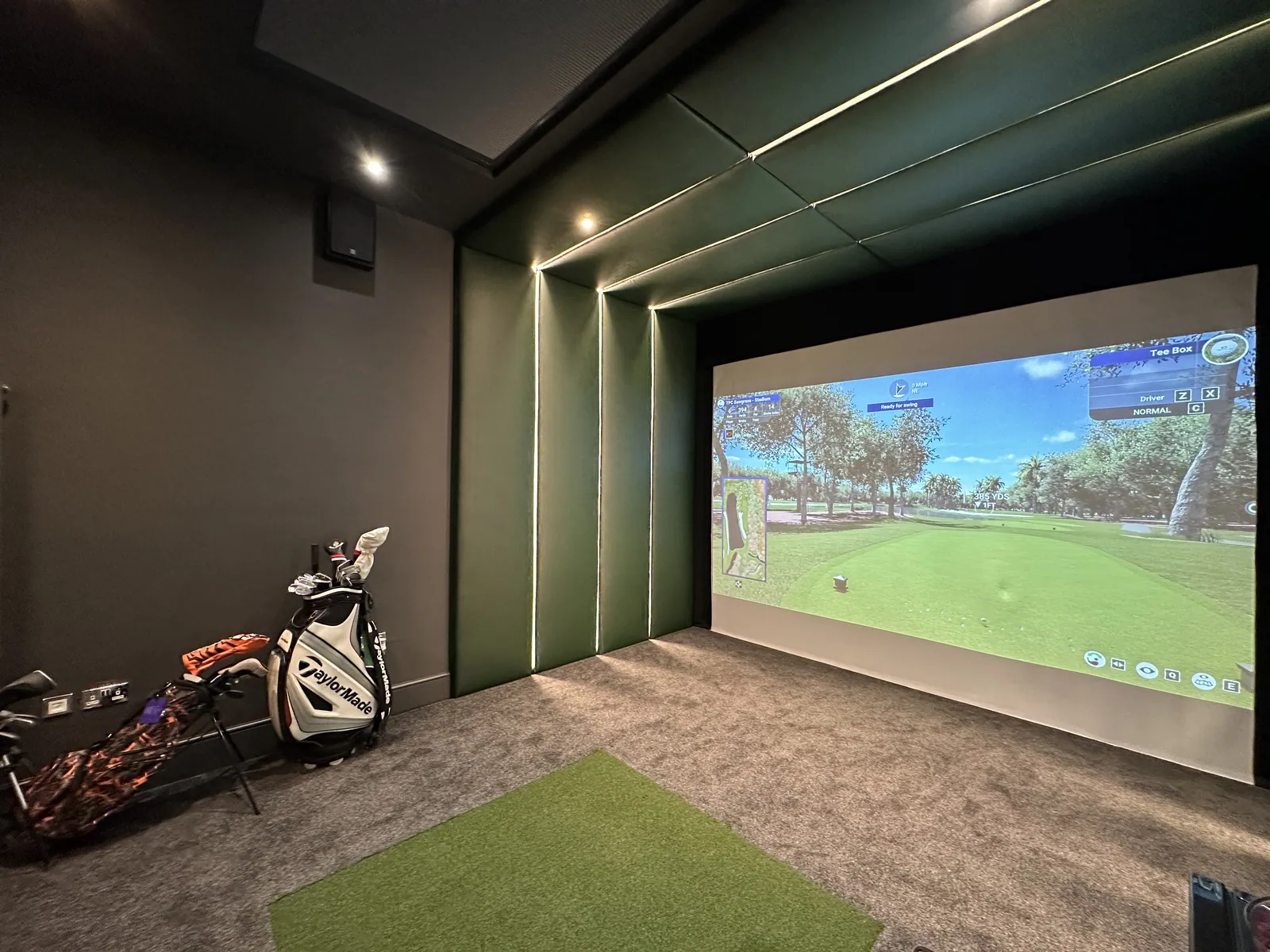 A golf simulator screen set in a lit enclosure, with a turf mat in front